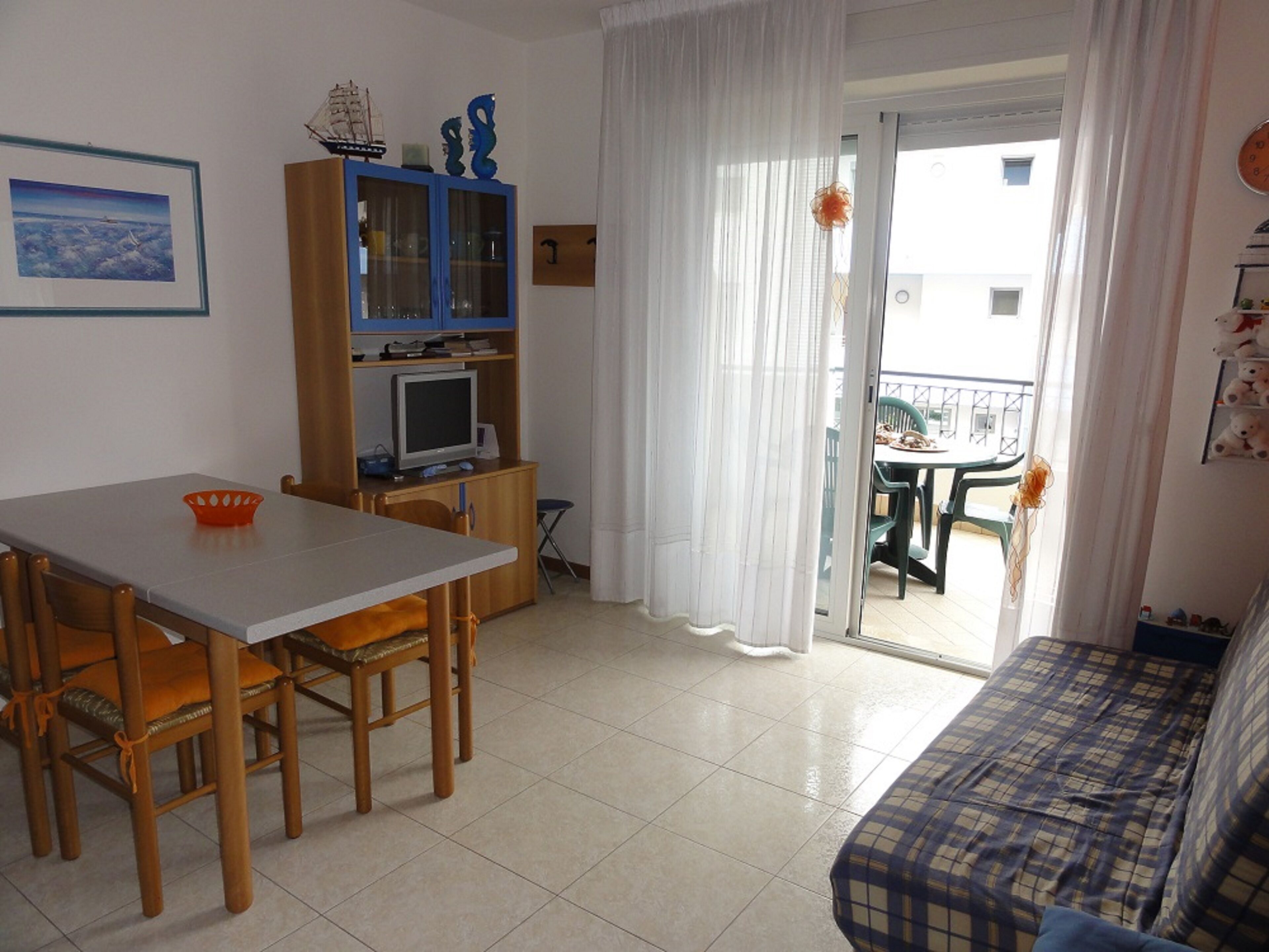A room with a dining table, chairs, a sofa, a TV, and a balcony with a view.