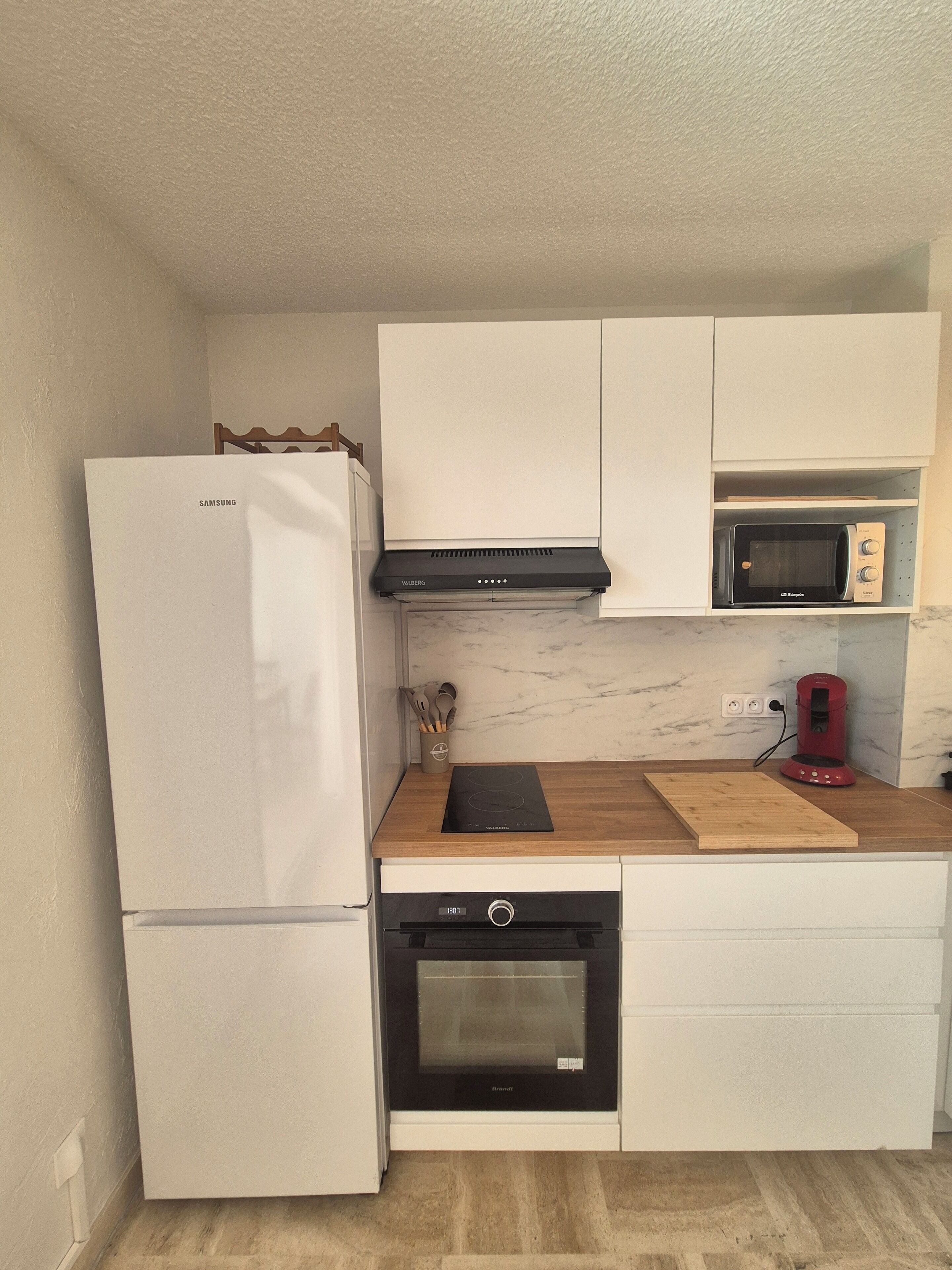 Een kleine keuken met een witte koelkast, een magnetron, een fornuis en een koffiezetapparaat.