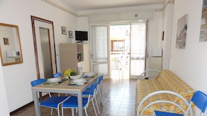 A dining area with a table set for four, blue chairs, a sofa with patterned cushions, and a television on a cabinet. - Bright flat close to the spa (Bibione)