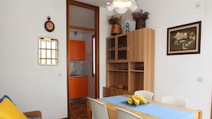 A dining area with a wooden table, white chairs, and a blue tablecloth. There is a kitchen in the background with orange cabinets. - Apartment in a shared villa with garden (Bibione)