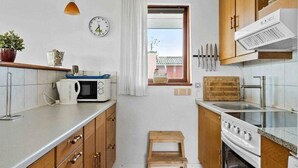 A compact kitchen with wooden cabinets, a microwave, a stove, and a small window with a view. - 5 Star Holiday Home in Bogense (Bogense)