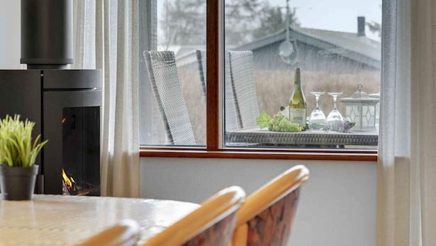 A cozy room with a fireplace, a dining table, and a view of a house through a window. - 5 Star Holiday Home in Bogense (Bogense)