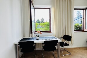 Une pièce avec un bureau blanc, deux chaises noires, une fenêtre donnant sur un paysage verdoyant et une structure bleue, ainsi qu’un radiateur placé sous la fenêtre.