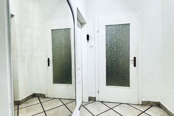 Un pasillo con dos puertas blancas, cada una con un panel de vidrio texturizado y un pomo negro. El suelo está cubierto de baldosas con un diseño sencillo.