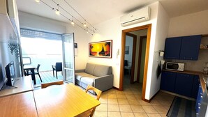 A compact living space with a dining area, a sofa, a kitchen with blue cabinets, and an air conditioning unit. - Bibione apartment with pool and solarium access (Bibione)