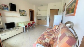 A compact living space with a sofa, a TV, a dining table with chairs, a kitchen area, and a refrigerator.