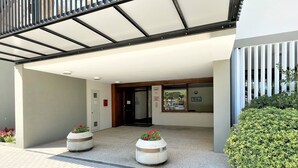 Una entrada de edificio con un toldo, dos macetas con flores y un seto verde.