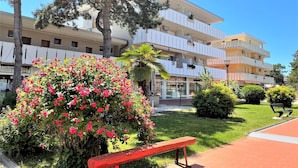 A multi-story hotel with balconies, a garden with pink flowers, and a red bench.