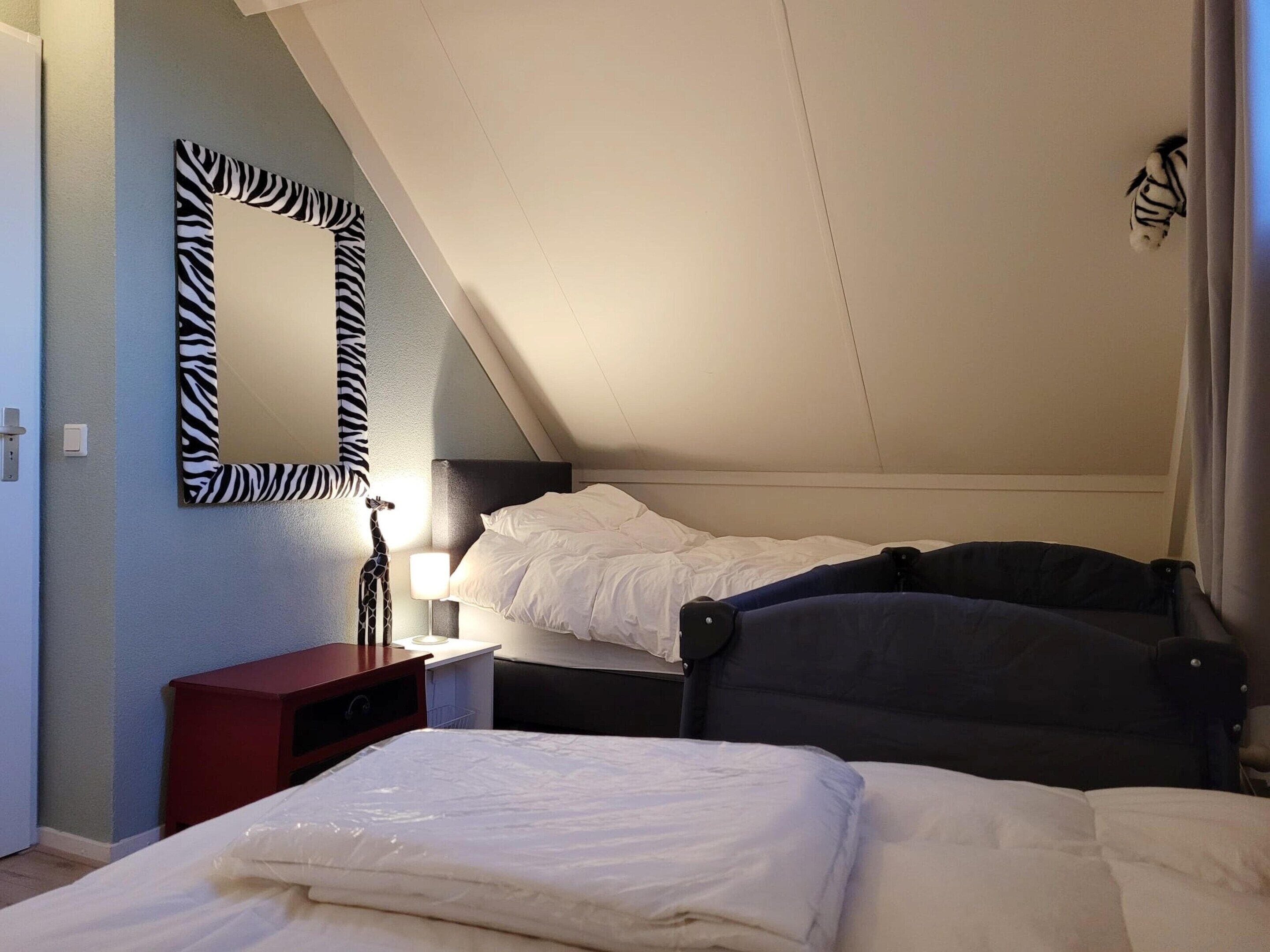 Ein Schlafzimmer mit schrägem Dach, einem zebraartig gestreiften Spiegel, einem Bett mit weißer Bettwäsche, einem Nachttisch mit Lampe und einem roten Beistelltisch.