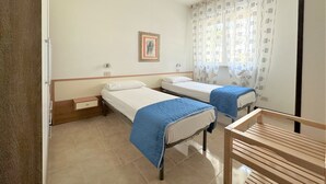 A room with two single beds, a wooden chair, a small table, a framed picture on the wall, and a window with patterned curtains. - Bibione apartment close to promenade (Bibione)