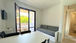 A compact room with a dining table, a gray sofa, and a sliding glass door leading to an outdoor balcony. - Bibione flat with pool access (Bibione)