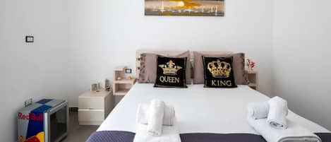 A hotel room with a bed, two pillows with 'QUEEN' and 'KING' designs, a painting of a sunset, a Red Bull cooler, and a small table with decorative items.