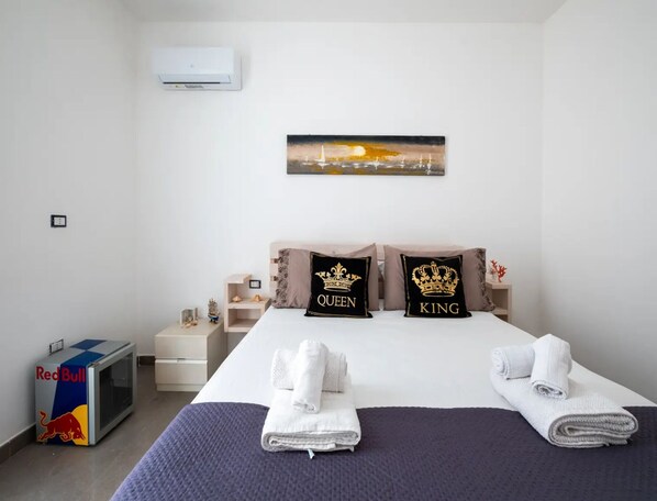 Une chambre d’hôtel avec un lit, deux oreillers ornés des motifs « QUEEN » et « KING », un tableau représentant un coucher de soleil, un réfrigérateur contenant des boissons Red Bull et une petite table décorative.