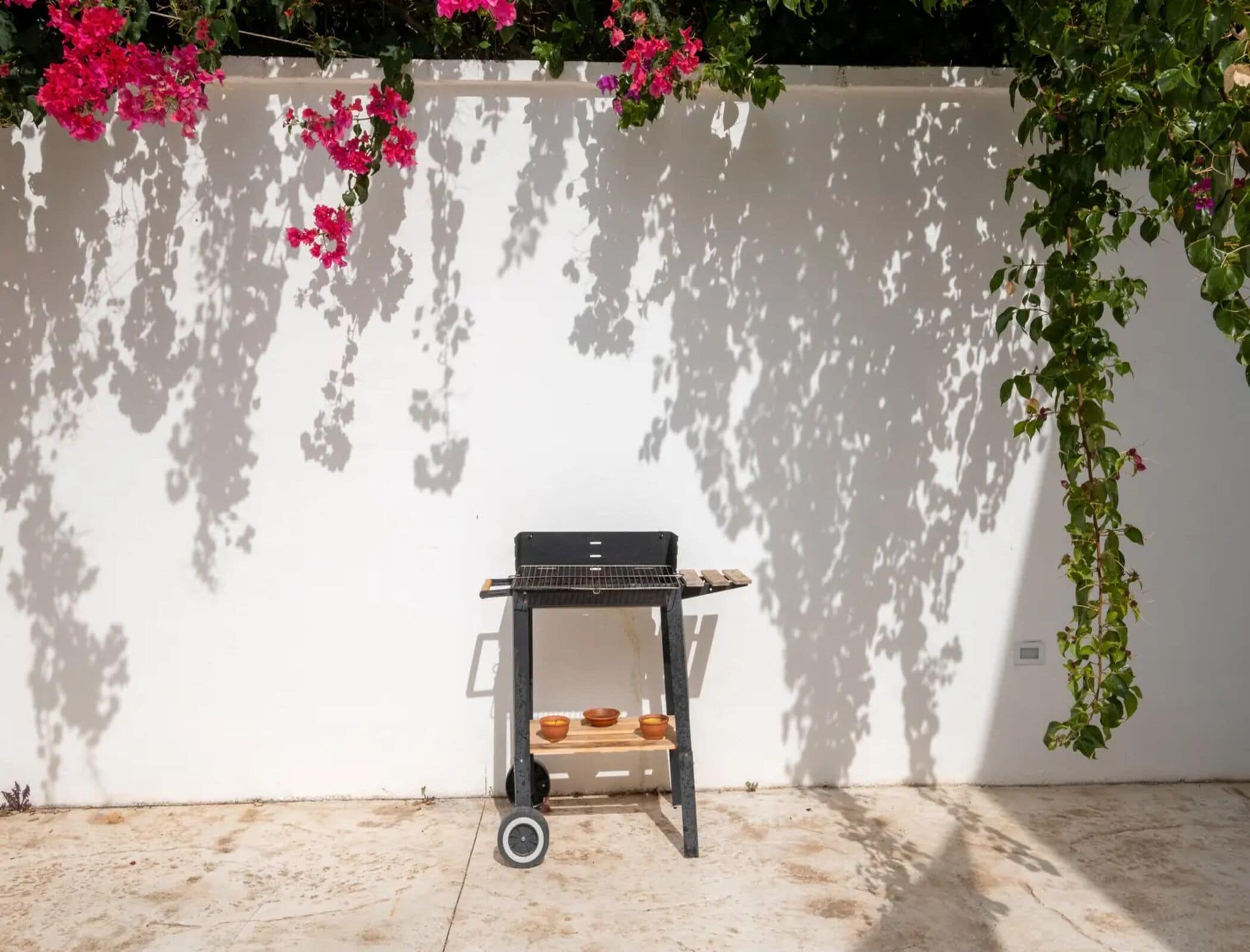 A patio area with a barbecue grill on a stand, a small shelf with bowls, and a concrete floor.
