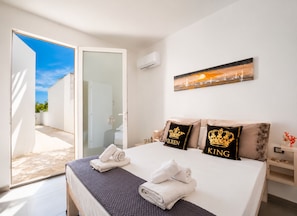 Une chambre à coucher avec un lit, deux oreillers ornés des motifs « QUEEN » et « KING », un tableau et une porte donnant sur un balcon.