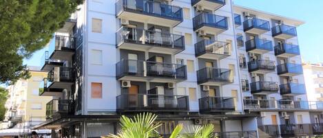 A multi-story residential building with balconies, air conditioning units, and a small outdoor seating area with chairs and a table.