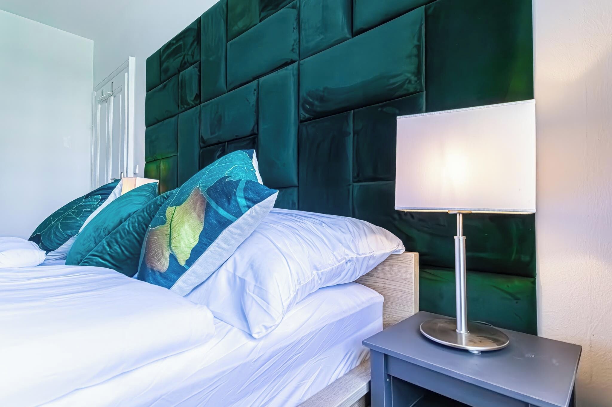 Una cama bien hecha con sábanas blancas y almohadas decorativas, un cabecero tapizado verde moderno y una mesita de noche con lámpara.