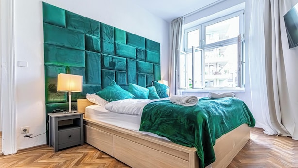 A modern bedroom with a large green headboard, a wooden bed frame, a nightstand with a lamp, and a window with a view of buildings. - 3BR Apt 15 Min from Landstraße Hub (Vienna)