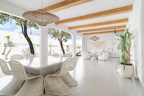 A spacious dining area with a white table and wicker chairs, a hanging light fixture, and a view of the outdoors. - Casa Contenta Canggu (Canggu)