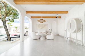 A modern, minimalist room with a pool view, featuring a white sofa set, a hanging light fixture, and a large arched mirror. - Casa Contenta Canggu (Canggu)