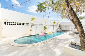 A swimming pool with a curved edge, surrounded by a white wall with decorative circular cutouts. There are potted plants and a tree with yellow flowers.