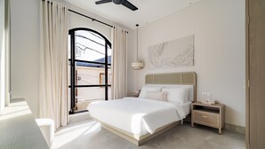 A modern hotel room with a large bed, a bedside table, a nightstand, a ceiling fan, and a view of the outside through a window with curtains. - Casa Contenta Canggu (Canggu)