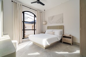 A modern hotel room with a large bed, a bedside table, a nightstand, a ceiling fan, and a view of the outside through a window with curtains.
