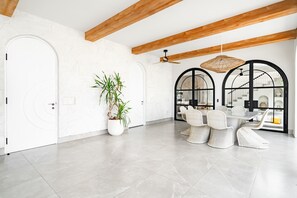 A spacious room with a modern design, featuring a white door, a potted plant, a dining area with wicker chairs, and a ceiling fan.
