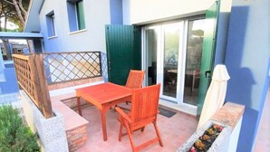 Eine Terrasse mit Tisch und Stühlen, ein kleines Blumenbeet und ein Gebäude mit blauer Fassade und einer Glastür.