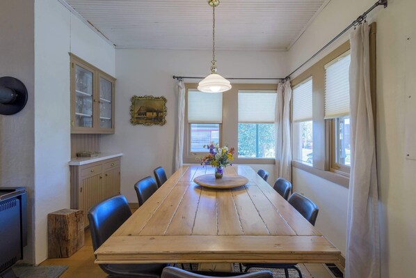 A dining area with a wooden table, chairs, a hanging light fixture, a cabinet, and a window with curtains.