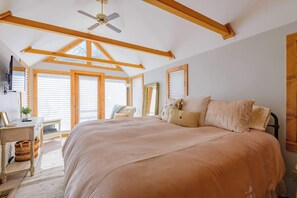 A bedroom with a large bed, wooden beams, a ceiling fan, and a view through large windows. - The Porch House, hot tub, pet friendly, historic! (Crested Butte)