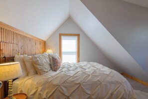 A bedroom with a sloped ceiling, a bed with a white duvet, a bedside lamp, and a window with blinds.