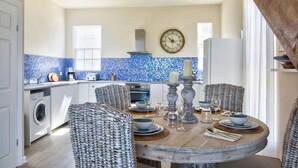 Une cuisine avec un mur recouvert de carreaux bleus, une horloge et un coin repas comprenant des chaises en osier et une table ronde.