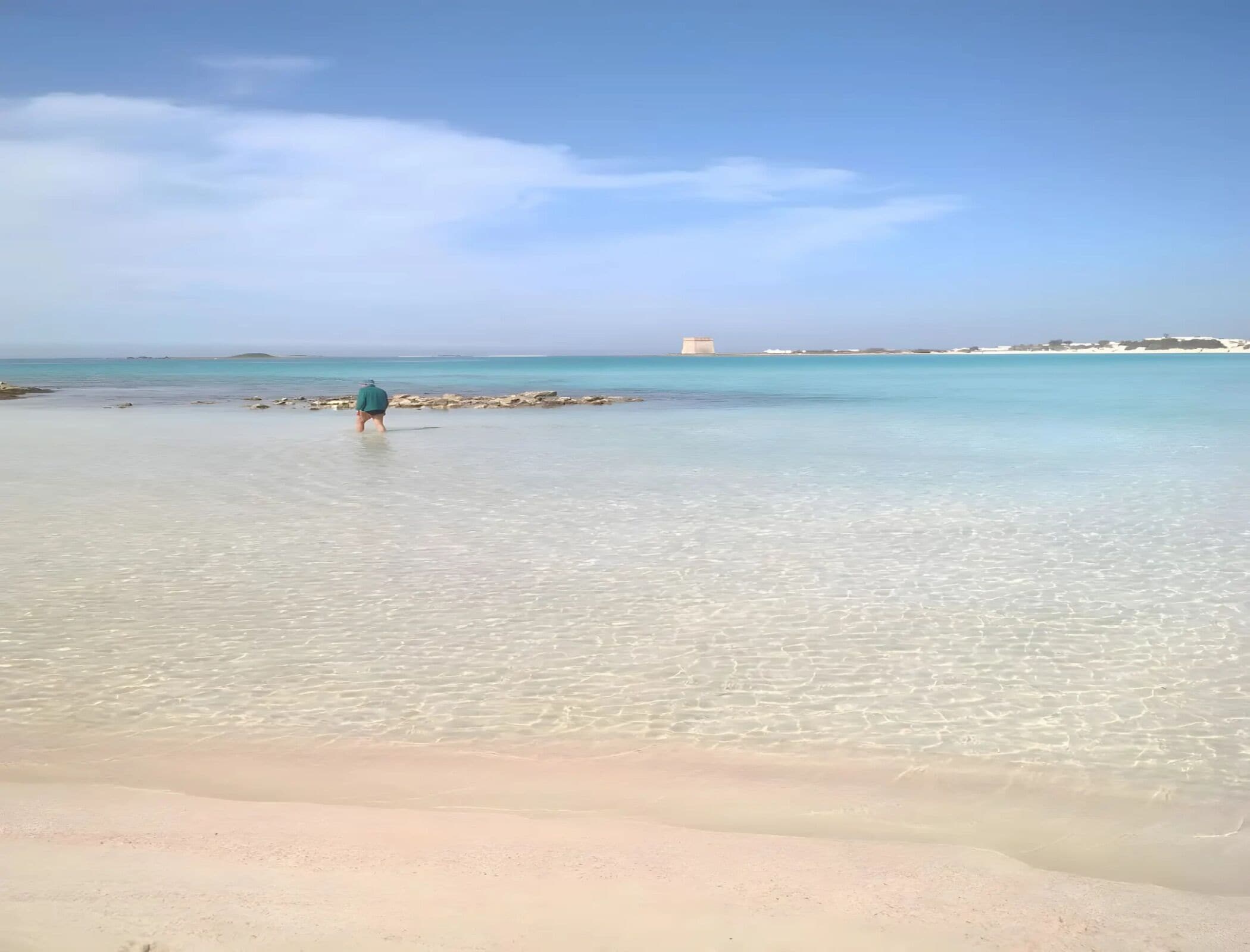 A beach with clear shallow waters and a person walking towards the water.