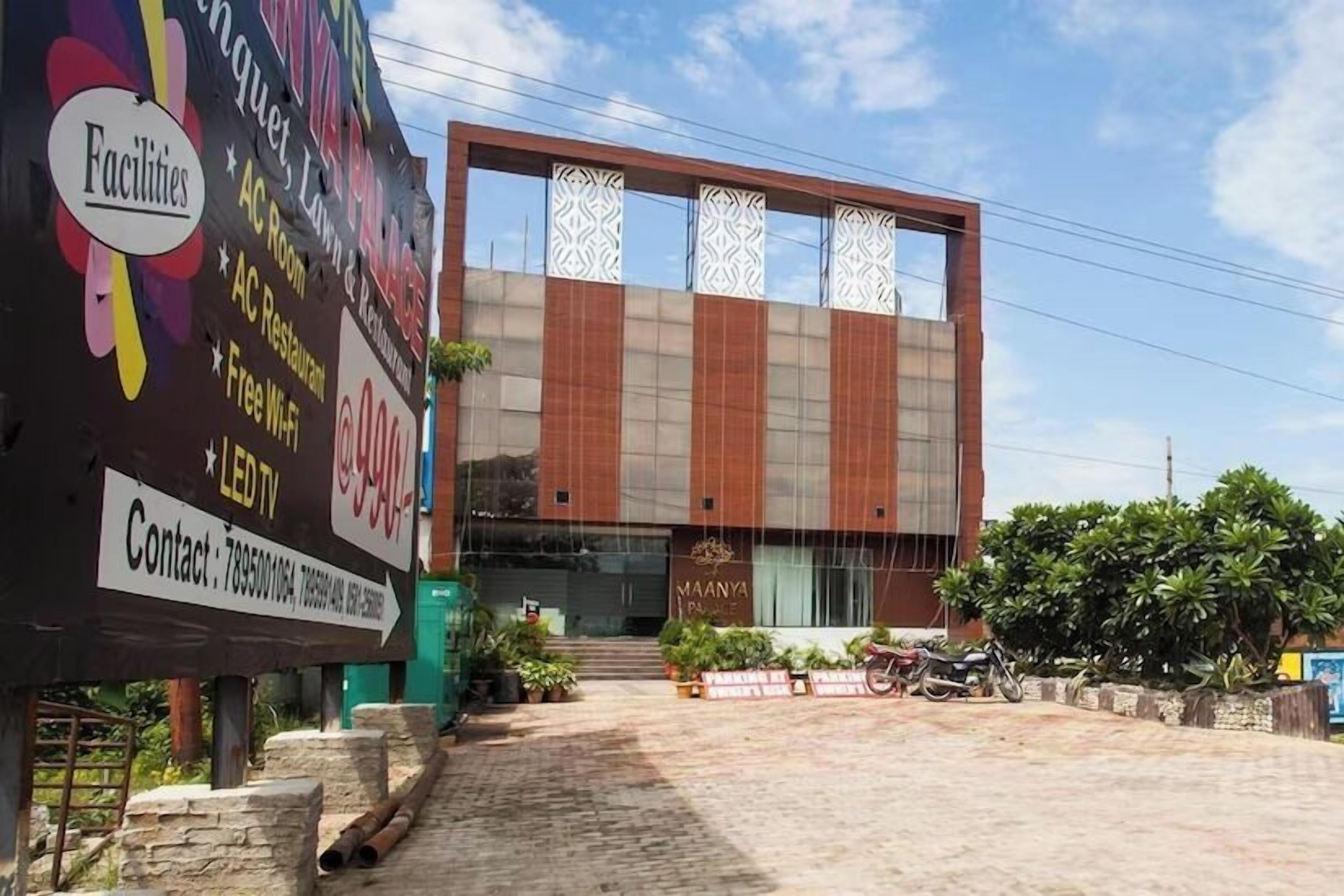 Un edificio moderno con un cartel que dice 'Maanya Palace' y un gran anuncio publicitario que muestra instalaciones como habitaciones con aire acondicionado, restaurante y Wi-Fi gratuito.