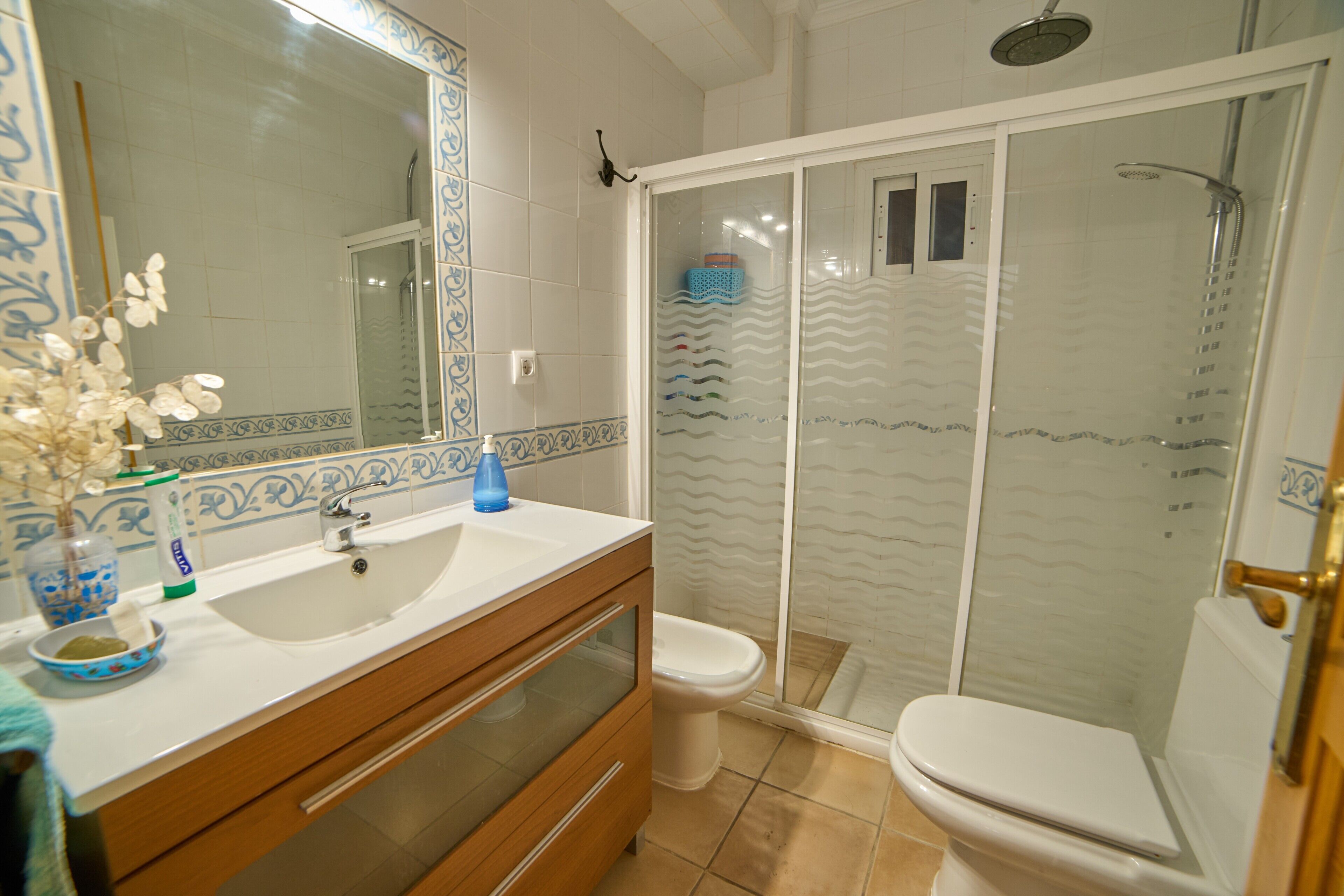 Un baño con lavamanos blanco, espejo y retrete, una ducha con puerta de vidrio y azulejos estampados.