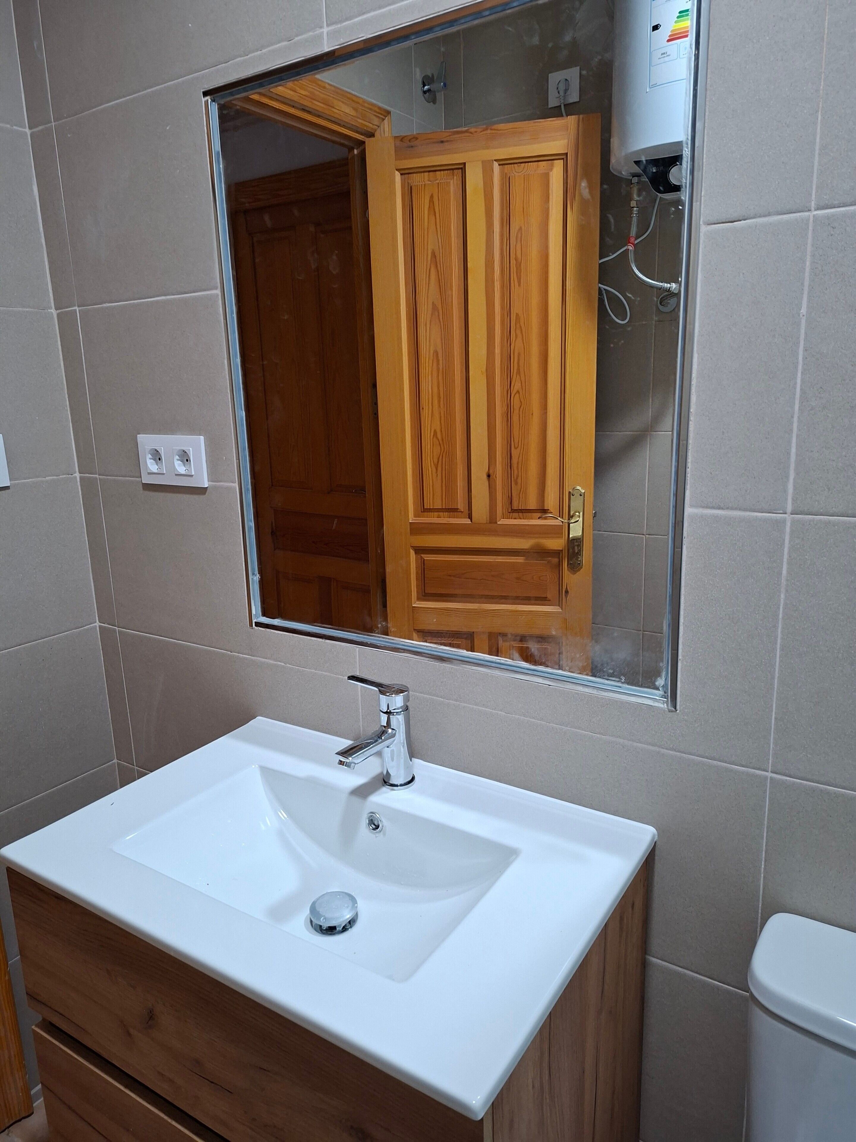A bathroom with a white sink, wooden cabinet, and a mirror reflecting a wooden door and a shower.