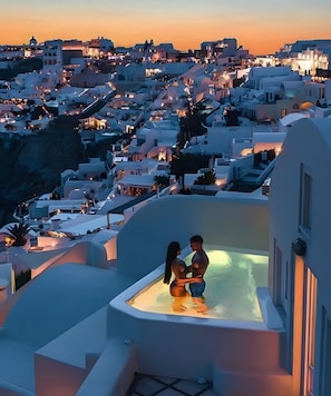 Un couple se détend dans un jacuzzi extérieur, l’eau est chaude, et ils ont une vue imprenable sur une ville aux maisons blanchies à la chaux, au moment du coucher du soleil.