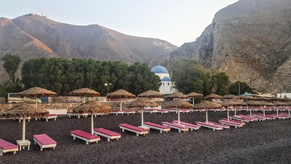 A beach with rows of sunbeds and thatched umbrellas, a white building with a blue dome, and mountains in the background.