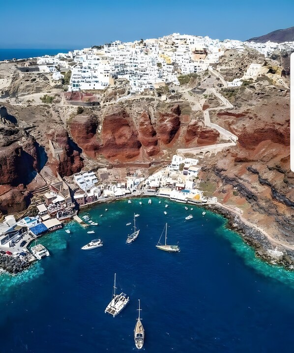 An aerial view of a coastal town with white buildings, a rocky coastline, and several boats anchored in a bay.