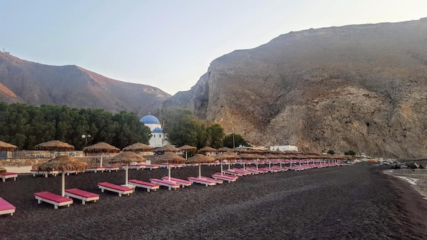 Une plage bordée de rangées de chaises longues et d’ombrelles en chaume, un bâtiment blanc surmonté d’un dôme bleu, et un paysage montagneux en arrière-plan.