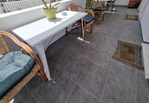 A balcony with a white table, wicker chairs, a potted plant, and a tiled floor.