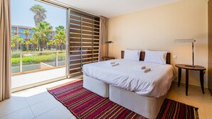 Ein Hotelzimmer mit einem großen Bett, einem kleinen Nachttisch, einem Fenster mit Blick auf Palmen und Gebäude sowie einem Balkon.