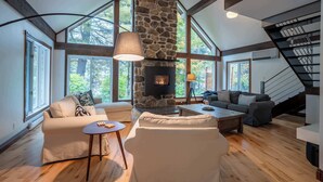 A spacious living room with a stone fireplace, large windows, and a staircase. - Lakefront | Quai 948 (Lac-Drolet)