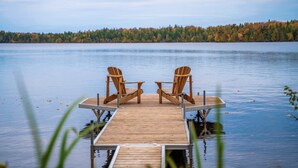 A wooden dock with two chairs facing each other, overlooking a calm lake with a forested shoreline. - Lakefront | Quai 948 (Lac-Drolet)
