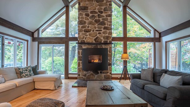 A cozy living room with a stone fireplace, large windows, and wooden flooring. - Lakefront | Quai 948 (Lac-Drolet)
