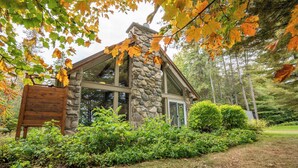 A stone cabin with large windows, surrounded by dense greenery and autumn leaves. - Lakefront | Quai 948 (Lac-Drolet)