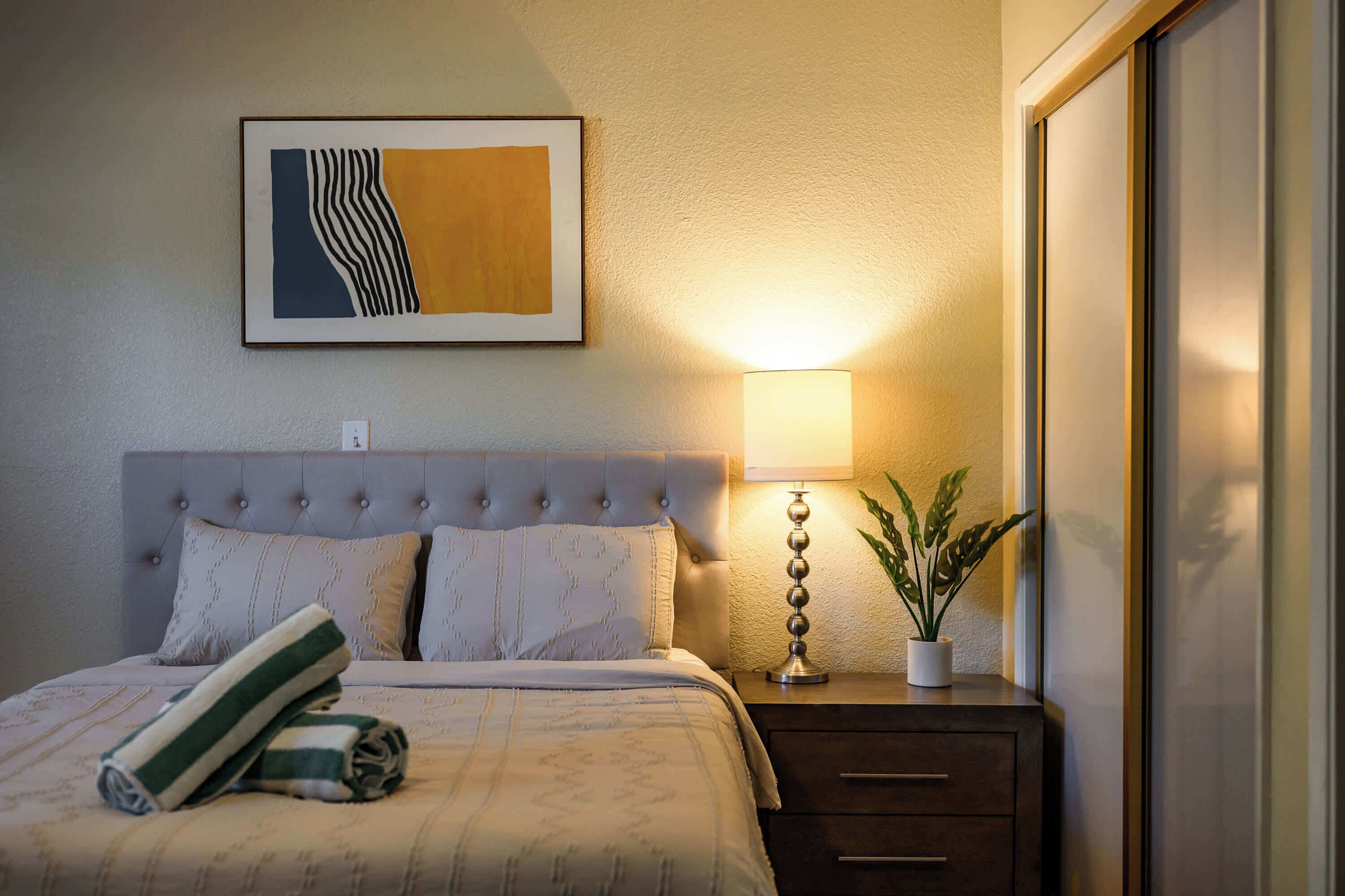 Een net opgemaakt bed met een gestreepte handdoek, een bedlamp, een ingelijst abstract schilderij en een potplant op een nachtkastje.