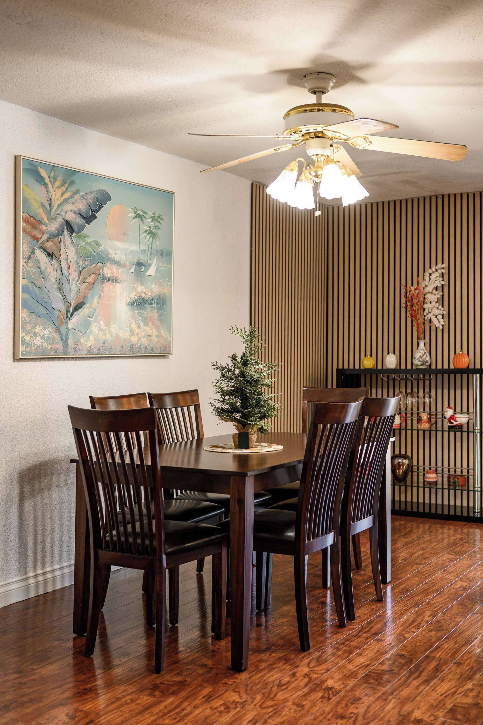 A dining area with a wooden table, six chairs, a ceiling fan, and a wall-mounted shelf with decorative items.