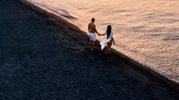 Een strandscène met een paar dat hand in hand loopt, een zonsondergang en boten in de verte.